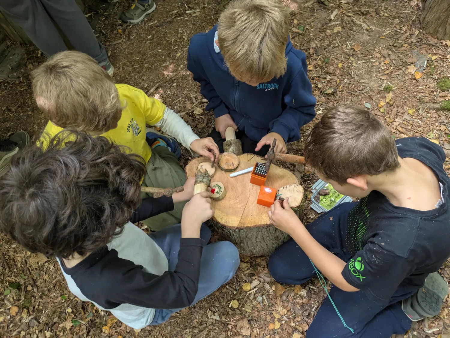 Four children sitting around a log stump, whittling and working with hand tools Four children sitting around a log stump, whittling and working with hand tools